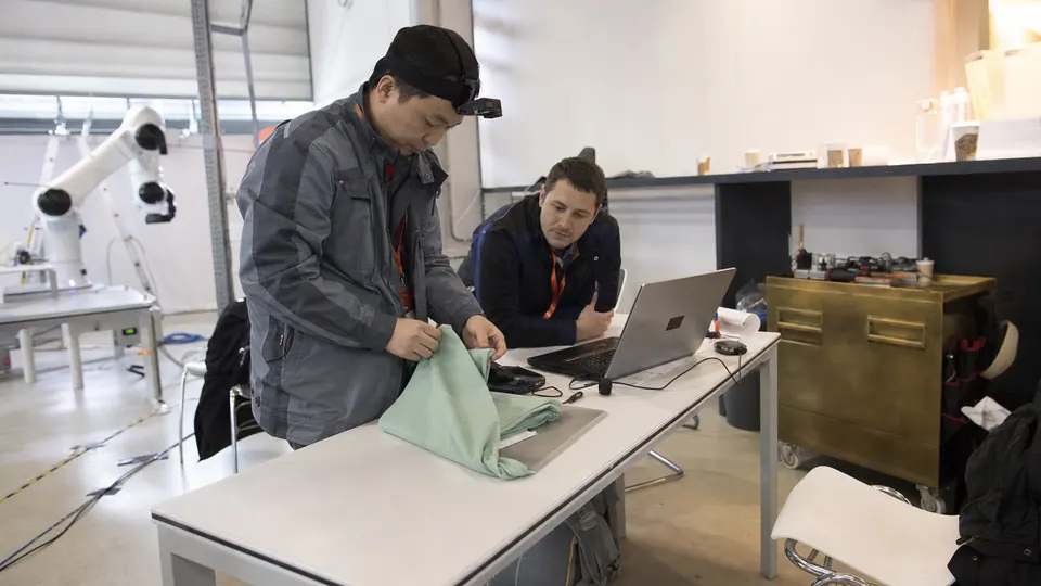 A worker wearing a head-mounted camera performs a hand task at a bench in a training hall while a colleague watches and an idle robot arm sits nearby.