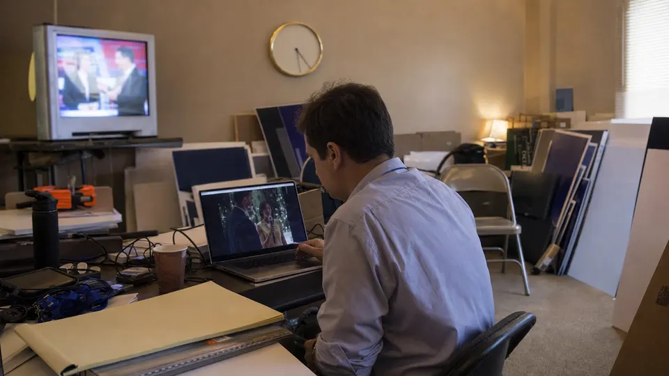 A campaign staffer reviews a video on a laptop in a cluttered campaign office with yard signs stacked behind, a brass clock on the wall and a muted TV in the corner.