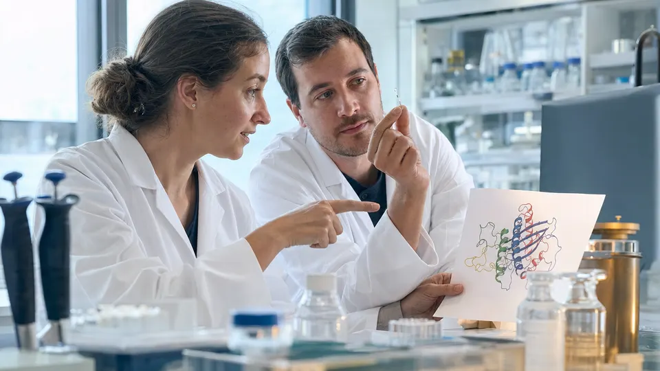 Two researchers in a protein crystallography lab examine a small crystal sample beside a ribbon-diagram model of a protein.