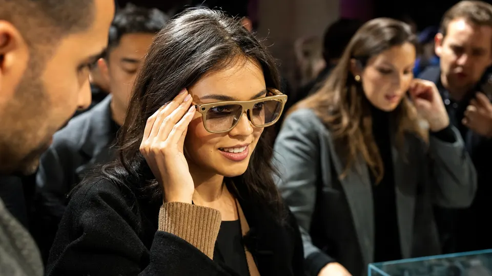 A smiling woman tries on gold-framed smart glasses at a product launch while other attendees gather around a display table.