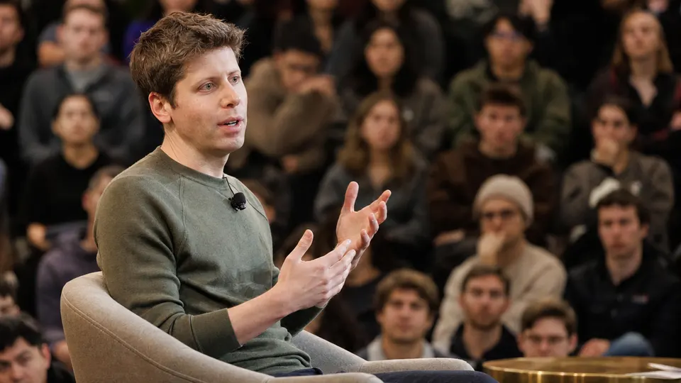 Sam Altman speaking in an onstage conversation in a university auditorium, with an audience blurred in the background.