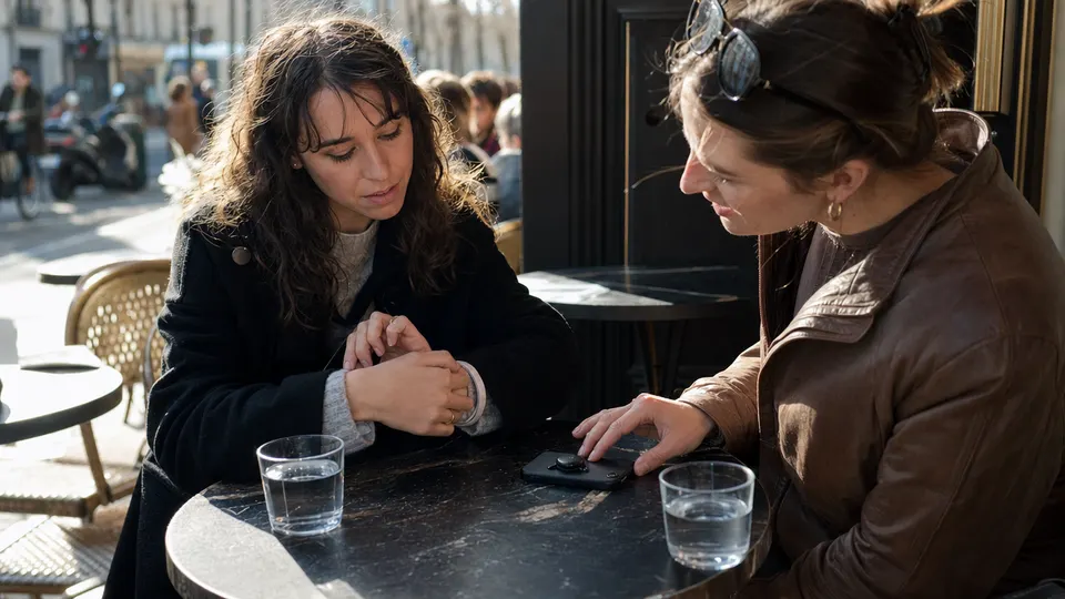 Two friends talk at a cafe table; one wears a wristband recorder and the other's phone has a small magnetic recorder attached to its back.
