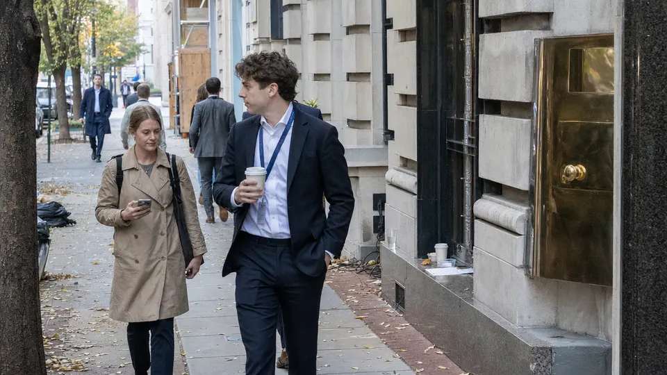 A professional with a lanyard and coffee walks past office buildings on an autumn street in Washington, DC, a brass door plate glinting beside the entrance.