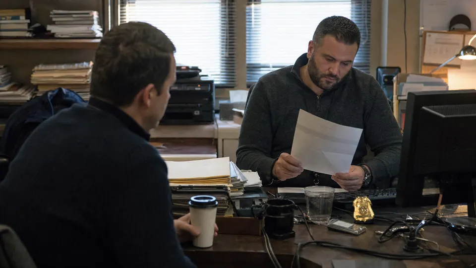 Two detectives review a printed email at a desk covered in case folders in a Philadelphia police office.