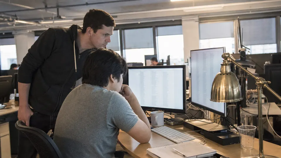 Two researchers in an office reviewing long columns of log text on dual monitors beside a brass desk lamp.