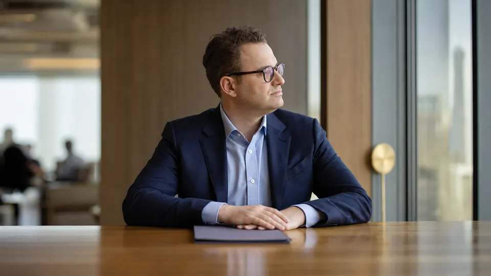 Illustrative photo of Dario Amodei seated at a conference table in a bright office, looking toward a window.