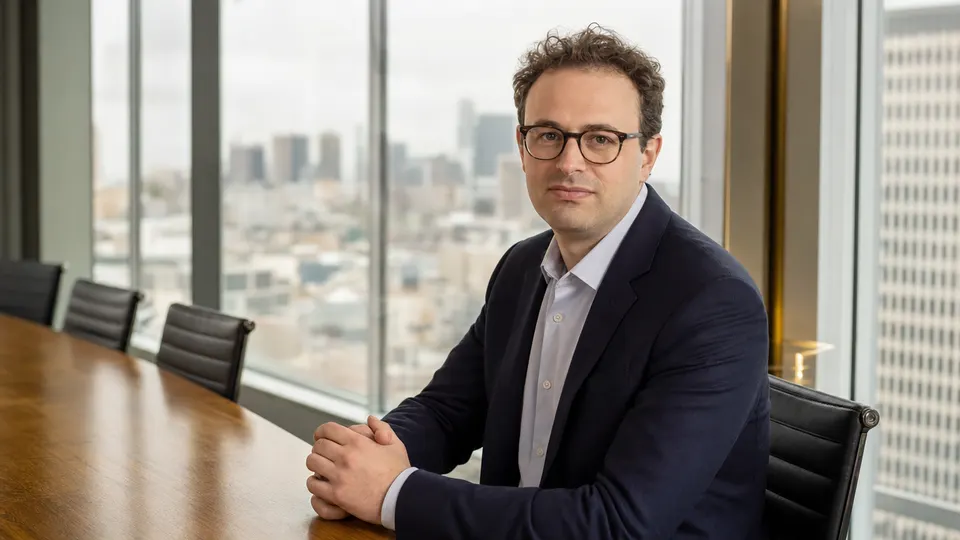 Portrait-style photo of a technology executive seated at a conference table in a bright glass-walled meeting room, looking calmly toward the camera.