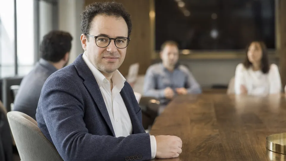 Dario Amodei seated at a conference table in a bright meeting room, looking toward the camera with colleagues softly blurred behind him (illustrative photo).
