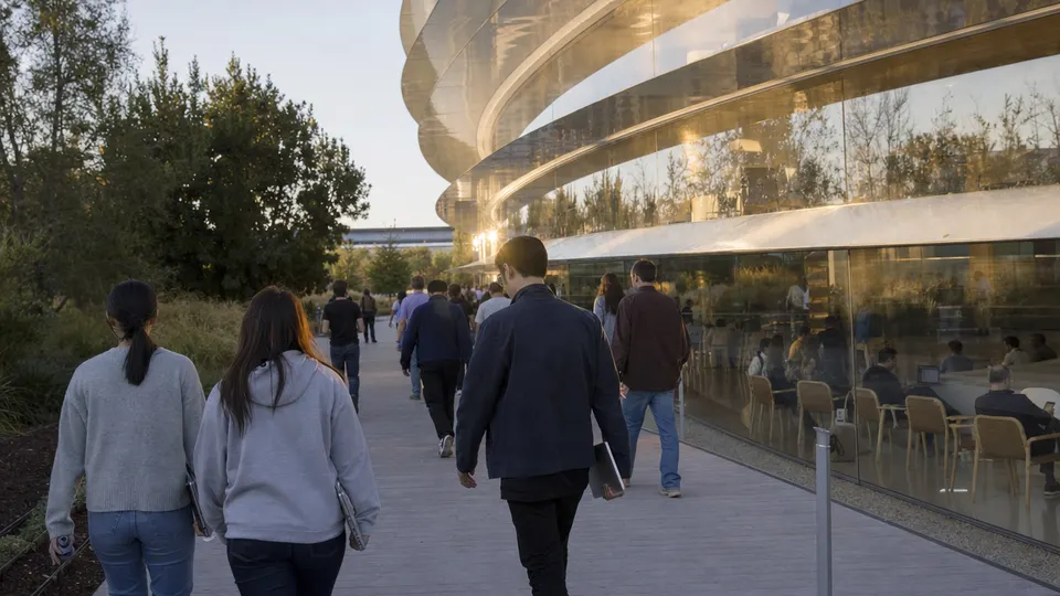 Apple employees walking along a path toward the circular Apple Park headquarters building in Cupertino in warm late-afternoon light.