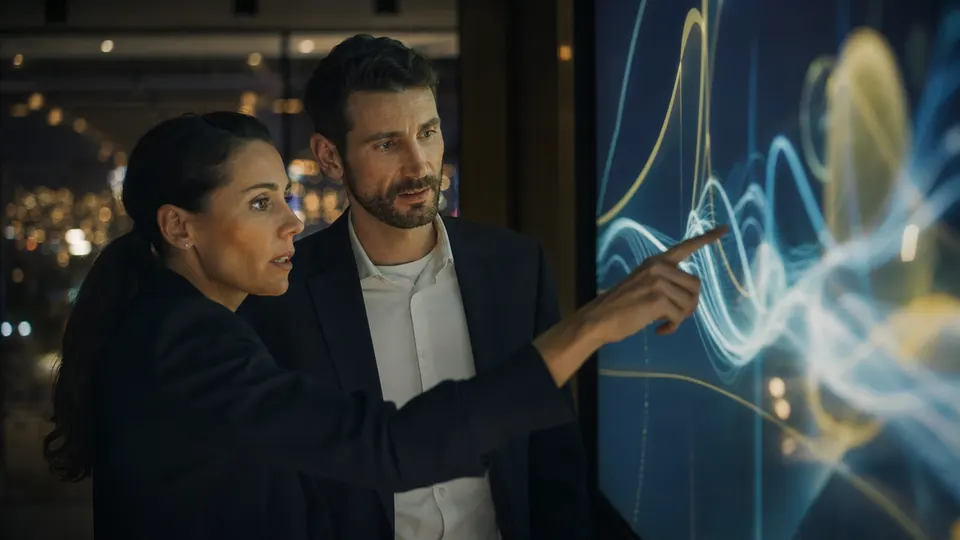 Two security analysts examine abstract data on a large screen in a dimly lit bank operations center.