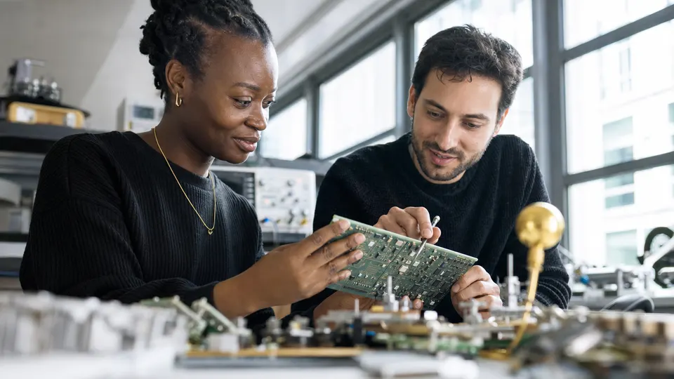 Two engineers examine a populated circuit board together at a lab workbench in soft daylight.