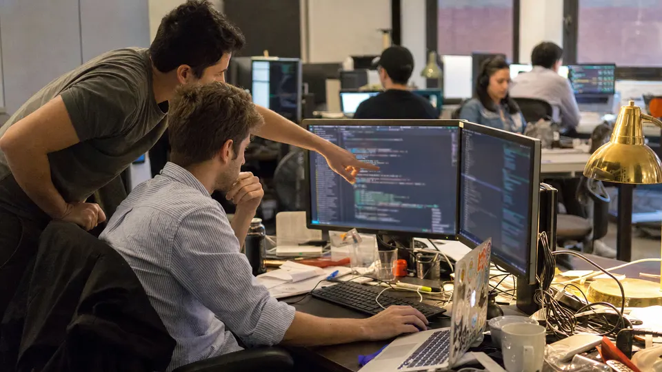 Two software developers review code on a monitor at a cluttered desk in an open-plan office, with a brass desk lamp glowing nearby.