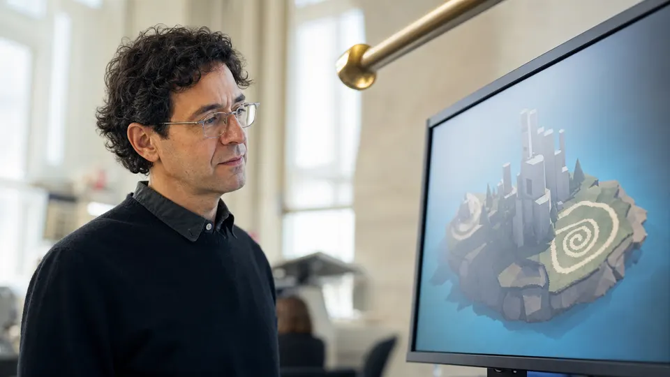 A researcher in a university lab studies a monitor showing a simple 3D island with blocky towers and a spiral pattern on the ground.