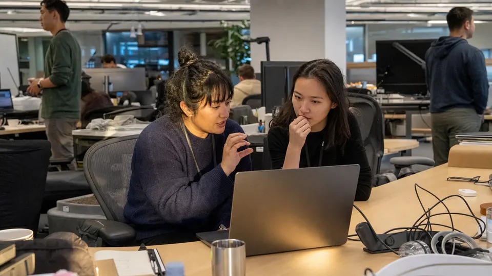 Two Anthropic employees discuss a laptop at the company’s San Francisco office, with late-afternoon sunlight on the desk.