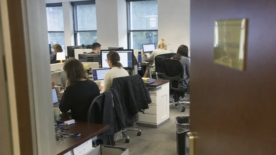 Government health agency staffers working at laptops in a busy open-plan Washington office, with a brass nameplate on a door in the background.