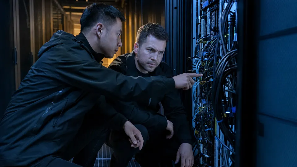 Two technicians inspecting an open rack of GPU servers in a large data center hall, lit by cool blue and warm amber light.