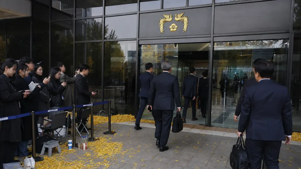 Government officials in dark suits enter the Government Complex Seoul as reporters record them, with golden ginkgo leaves on the plaza.