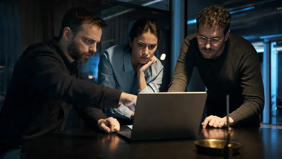 Three security engineers huddle around a laptop in a dim incident room, studying it intently.