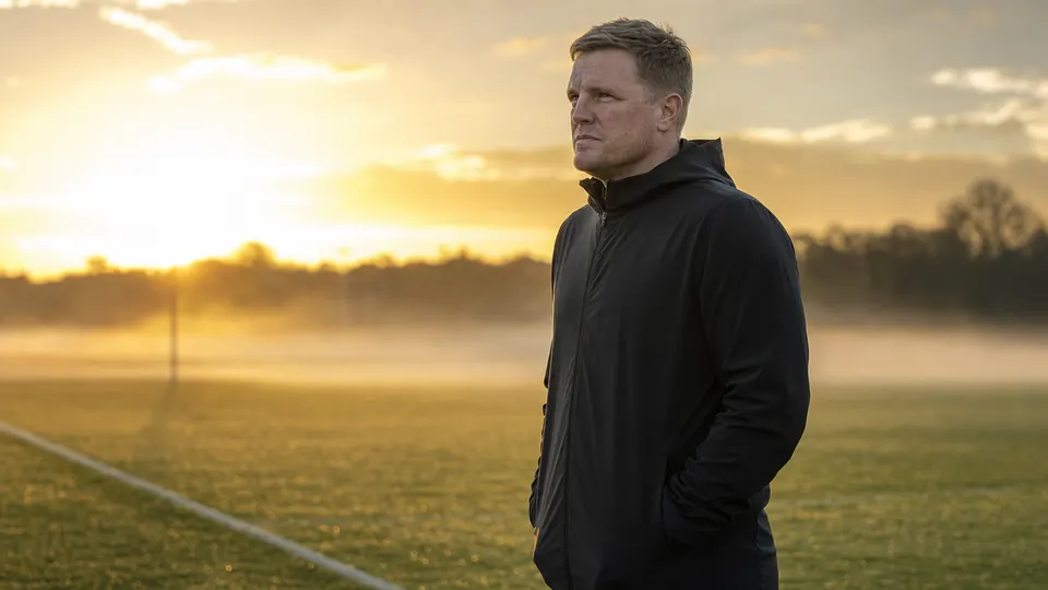 A manager in a plain dark training jacket stands at the edge of a misty training pitch at dawn, looking out across the grass.