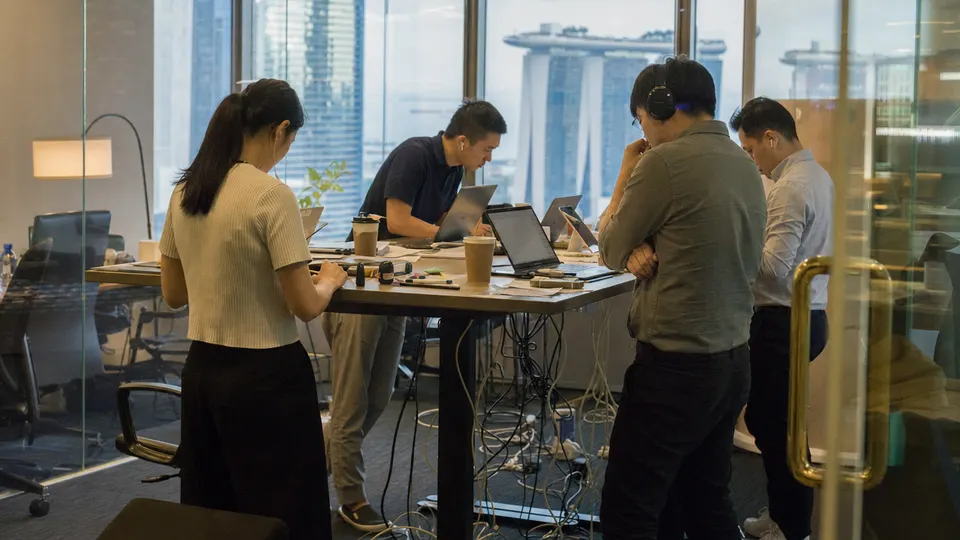 Employees gather around a standing desk with laptops and headsets in a Singapore office, with the city skyline visible through the window.