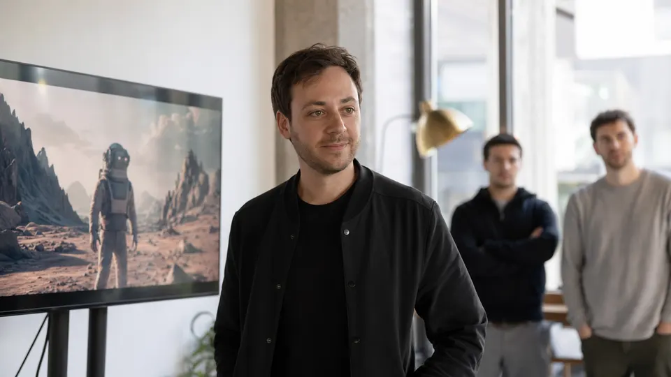 Portrait-style photo of a technology executive standing beside a monitor showing a still from a generated video, in a bright startup workspace.