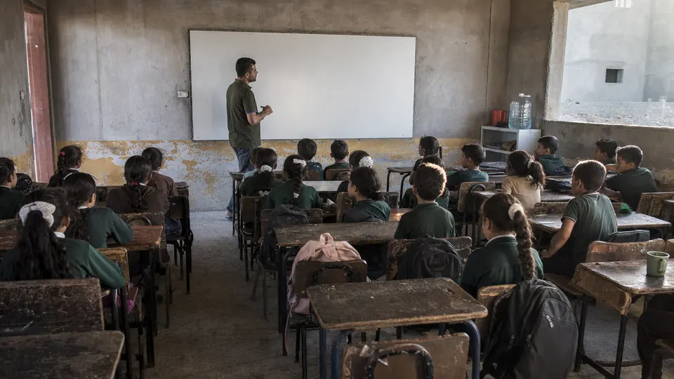 Children at wooden desks listen to a teacher at a whiteboard in a plain concrete classroom in Gaza, lit by afternoon sun.