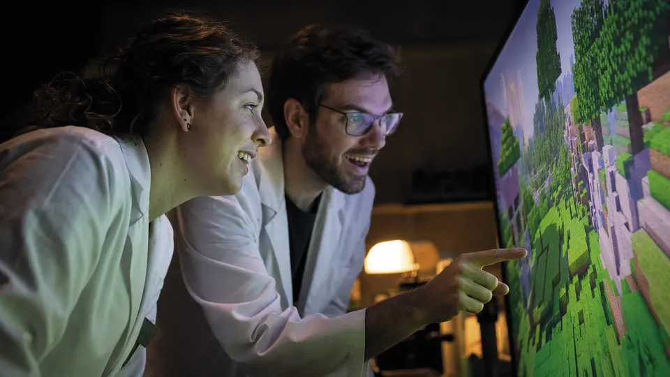 Two researchers in a dim lab react to a blocky video game world glowing on a large monitor.