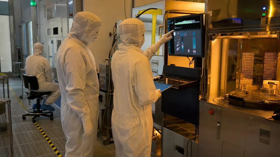 Engineers in cleanroom suits gather around wafer-handling equipment in a chip fabrication facility lit by amber light.