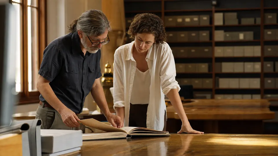 Two people examine a bound volume of official records at a table in an archive reading room.
