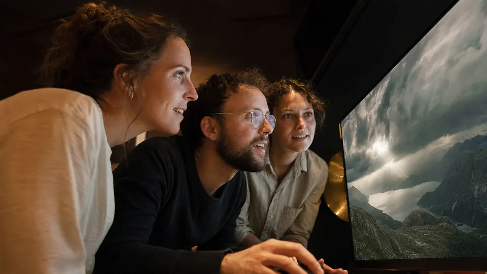 Researchers watch a generated landscape on a large screen change from sunshine to storm in a dim demo room.