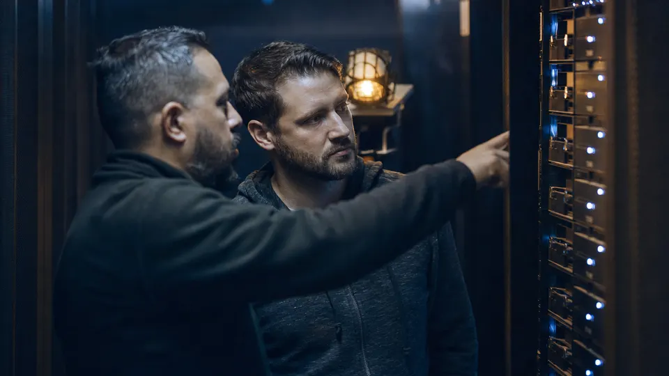Two security engineers confer in a dim server room lit by blue equipment lights, in an illustrative photo for a story on AI-driven hacking.