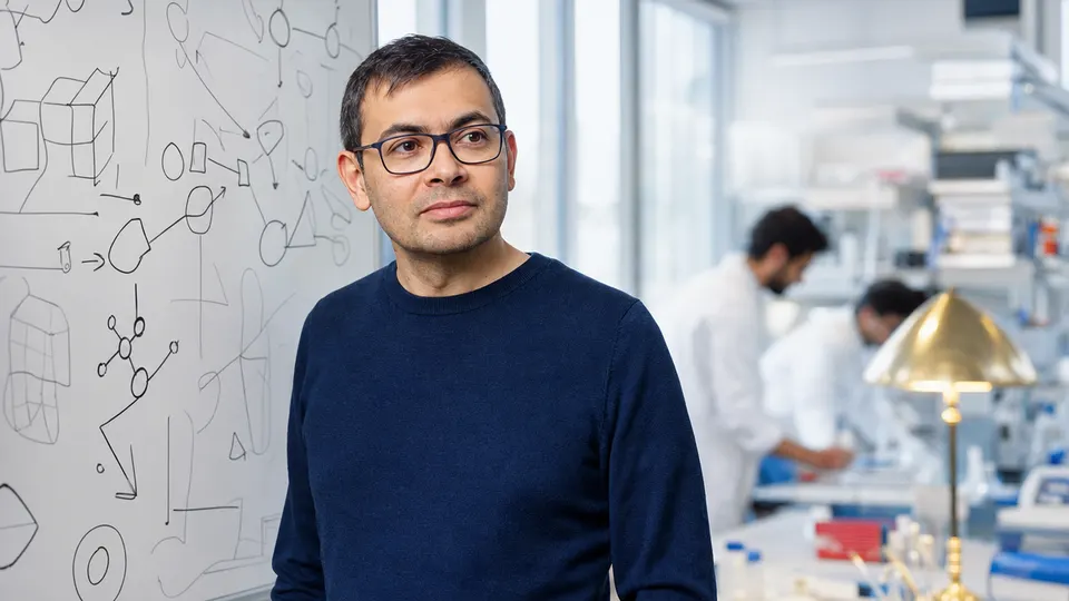 Illustrative photo of a man in a research lab standing in front of a whiteboard of diagrams, with researchers blurred behind him.
