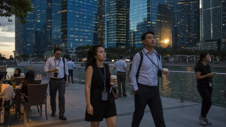 Commuters and office workers walk along the Marina Bay waterfront in Singapore at dusk, with glass towers catching golden light behind them.