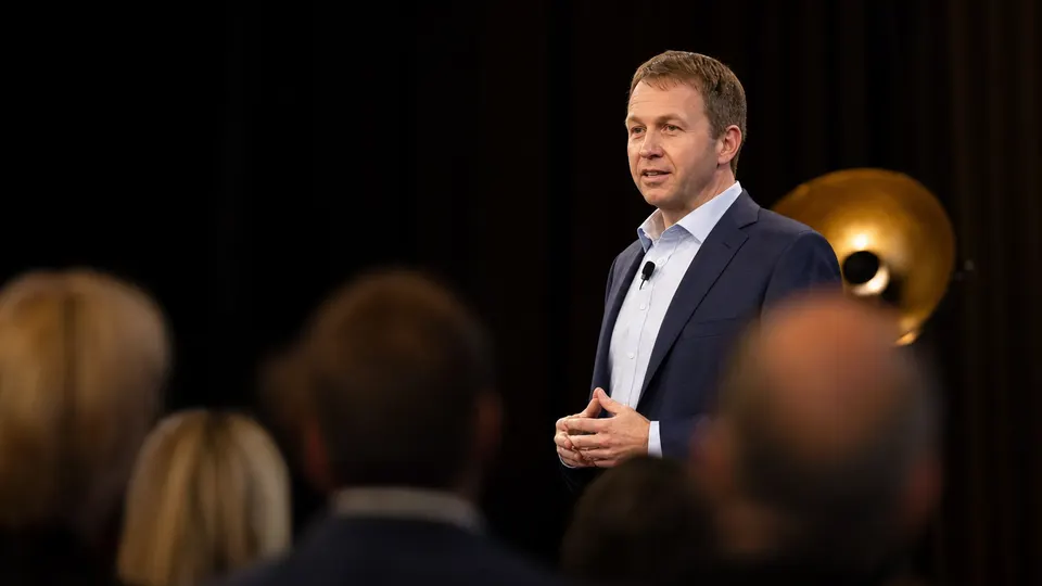 Illustrative photo of a technology chief executive speaking on stage at an investor event, lit by warm stage lights, with an audience blurred in the foreground.
