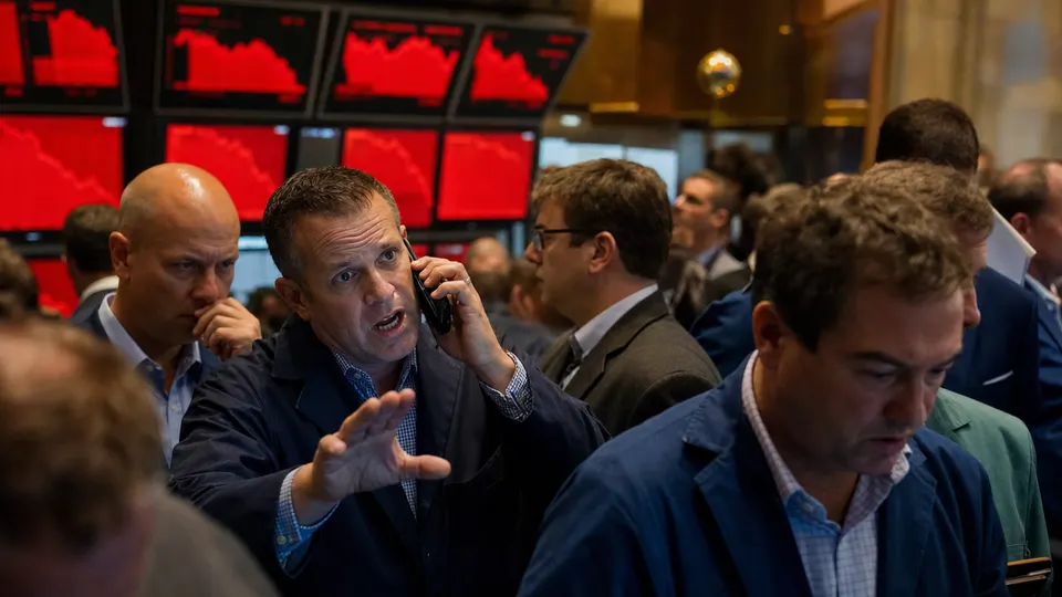 Traders on a stock exchange floor watch glowing red screens during a market selloff, one of them on a phone call.