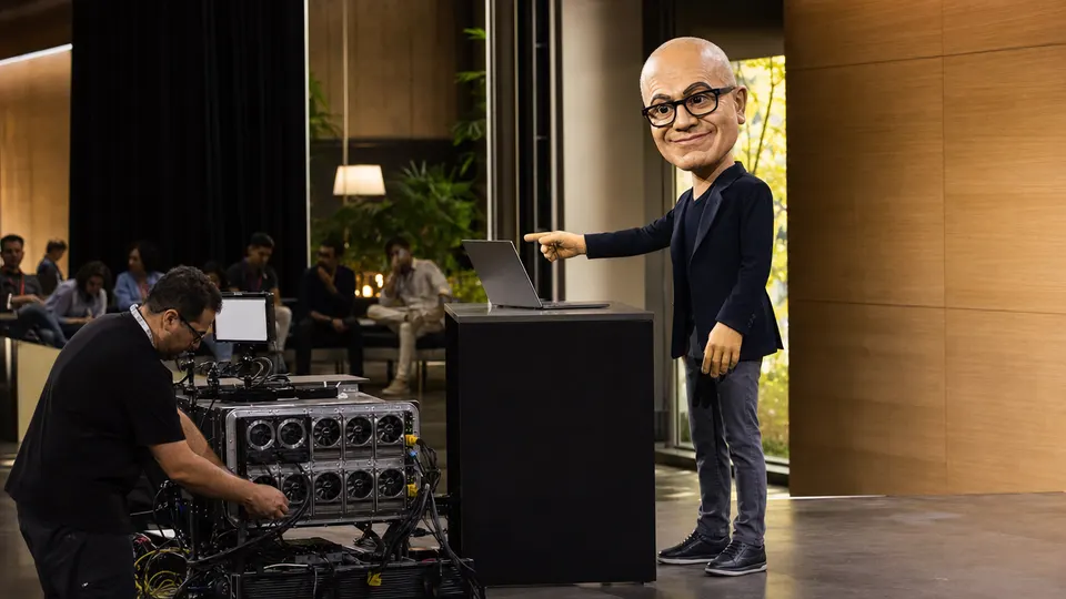 Satya Nadella puppet caricature on stage at a Microsoft campus auditorium, gesturing toward a Surface Laptop Ultra during the MAI-Code-1.1 Flash announcement, with a technician adjusting cables nearby and gold-tinted windows reflecting sunlight across the floor.