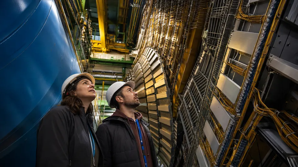 Two researchers in hard hats look up at a large particle detector inside an underground experimental hall.