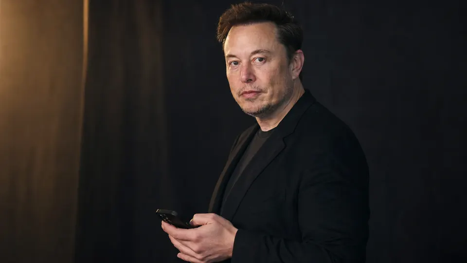 Elon Musk holding a smartphone, photographed in a dark studio in three-quarter view.