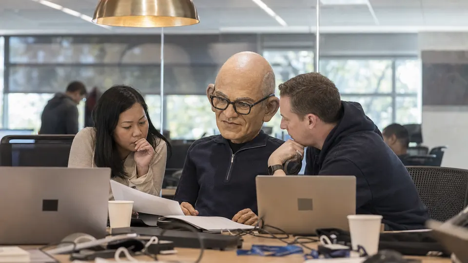 A puppet caricature of Satya Nadella reviews printed pages with colleagues at a table in a glass-walled meeting room, with laptops and coffee cups on the table and a brass pendant light overhead.