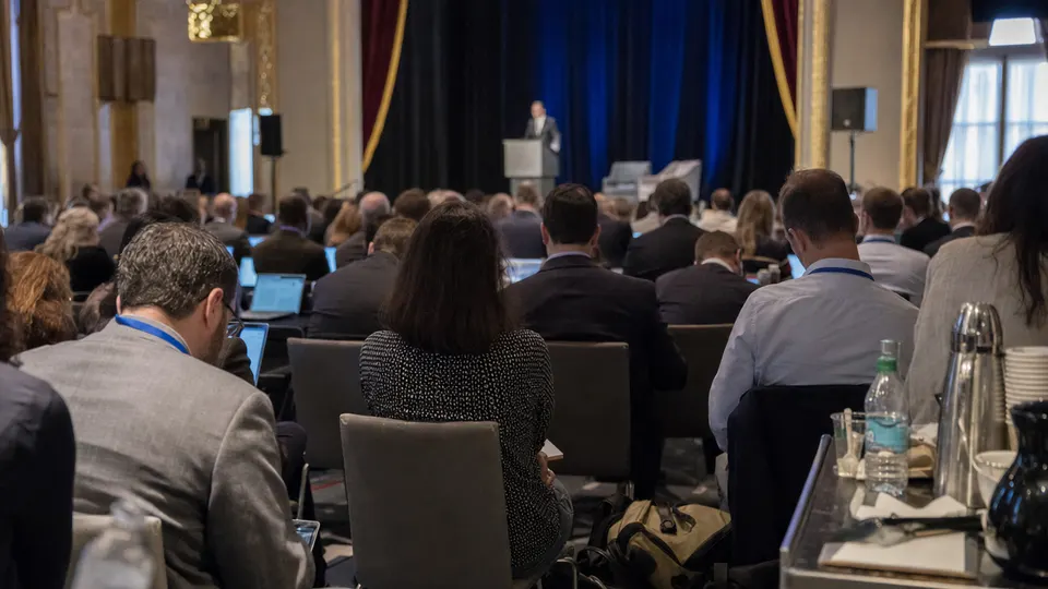 Attendees seated in a large conference hall in Washington, D.C. at a science summit, with a speaker at a podium and a gold-trimmed stage curtain behind.