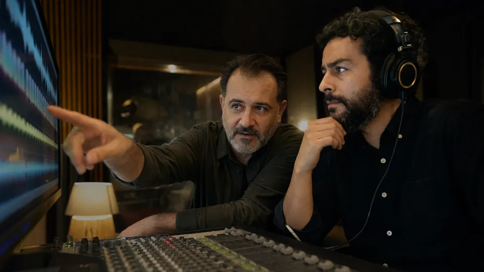 Two audio engineers in a dim recording studio listen to playback and study a waveform on a monitor.