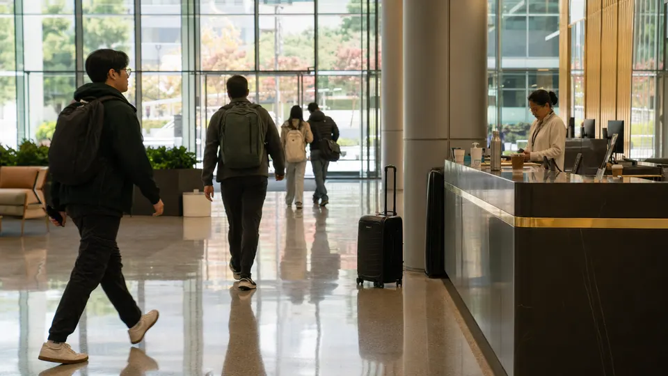 Employees with lanyards and coffee cups cross a bright glass-walled tech campus lobby with a gold-trimmed reception counter.