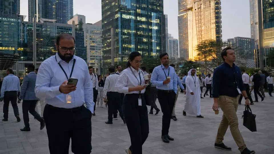 Office workers walk across a plaza at dusk in an Abu Dhabi business district, with glass towers behind them lit gold by the setting sun.