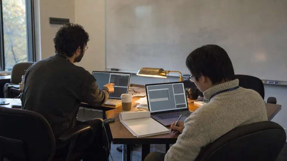 Two mathematicians review code on laptops at a seminar room table, with printed pages, coffee cups and a brass lamp.