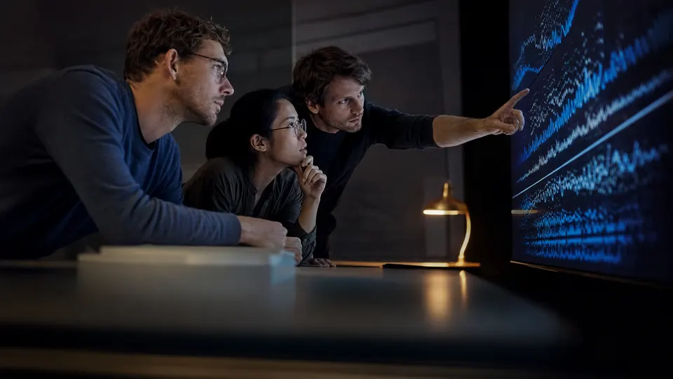 Researchers in a dim review room study abstract graphs on a wall display during a tense discussion.
