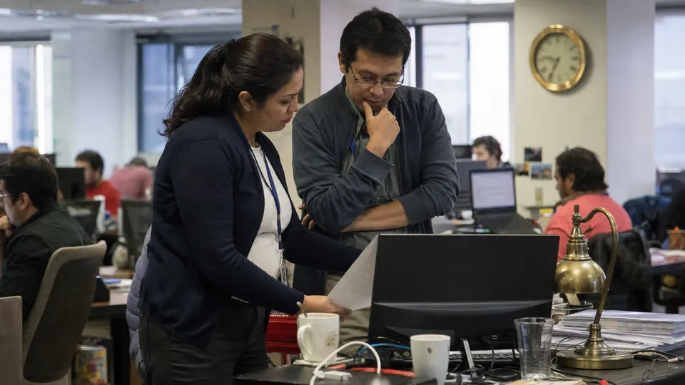 Two journalists review a printed document at a cluttered newsroom desk, lit by a brass lamp and a gold-framed wall clock.