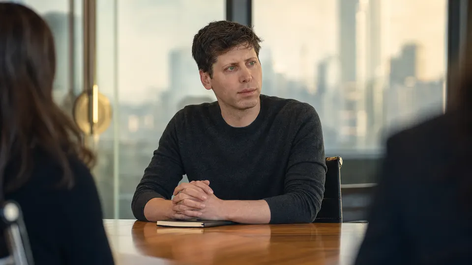 A man in a dark jacket sits at a conference table in a glass-walled meeting room, looking thoughtfully toward colleagues out of frame.