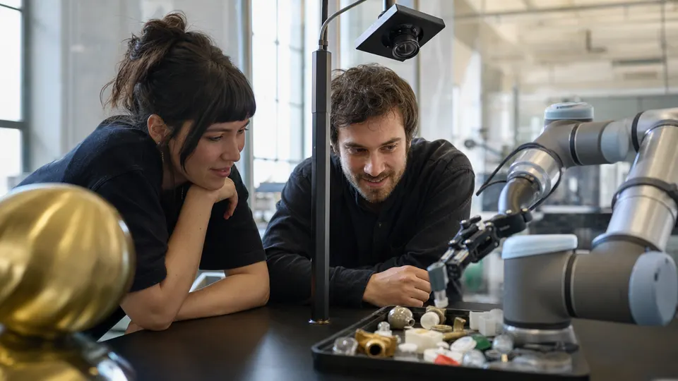 Two engineers watch a robotic arm sort small objects on a lab workbench under a camera rig.
