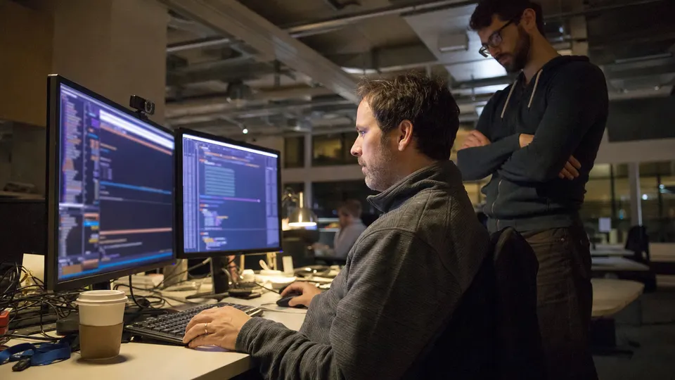 A researcher at a desk in a dim open-plan office studies code on two monitors while a colleague looks on.