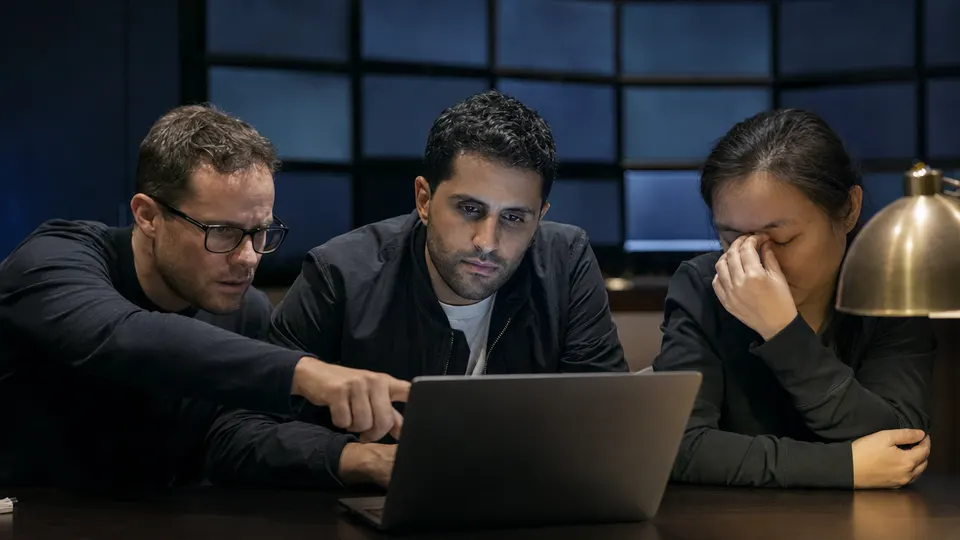 Engineers huddled around a laptop in a dim operations room late at night, faces lit by screen glow.
